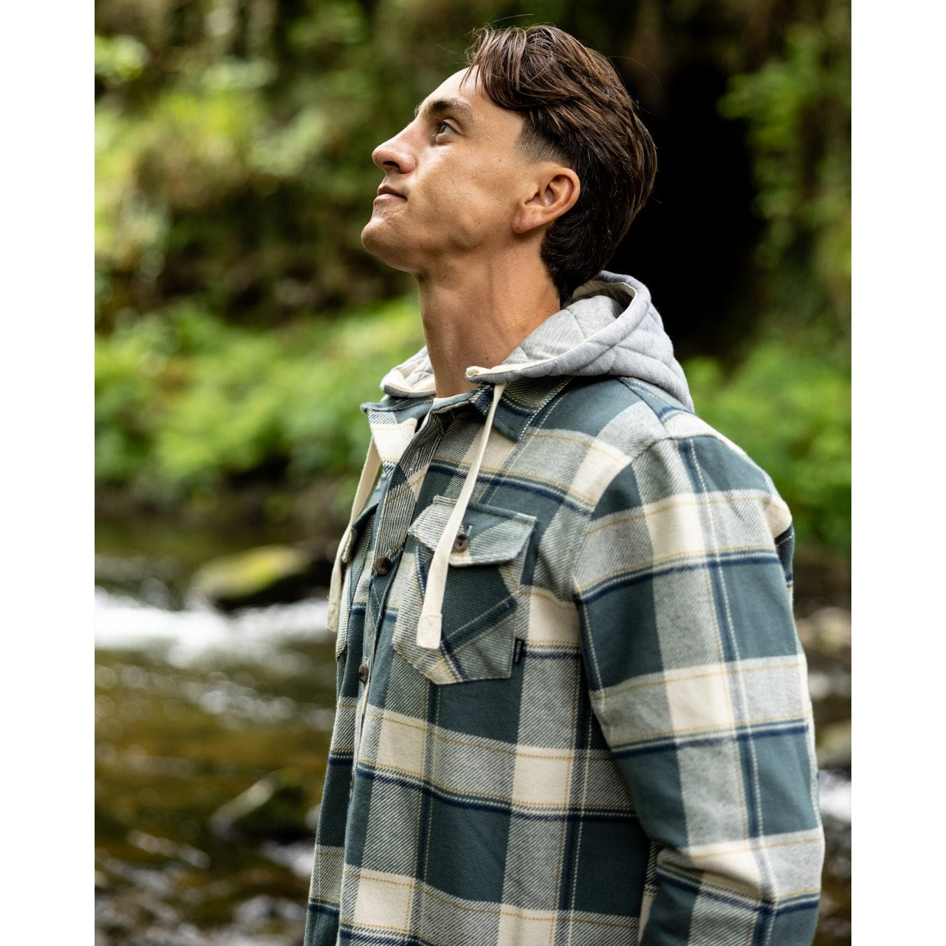 Side view of a man wearing a Saltrock Strait long sleeve check shirt in dark teal green with grey detachable hood and chest pockets outdoors