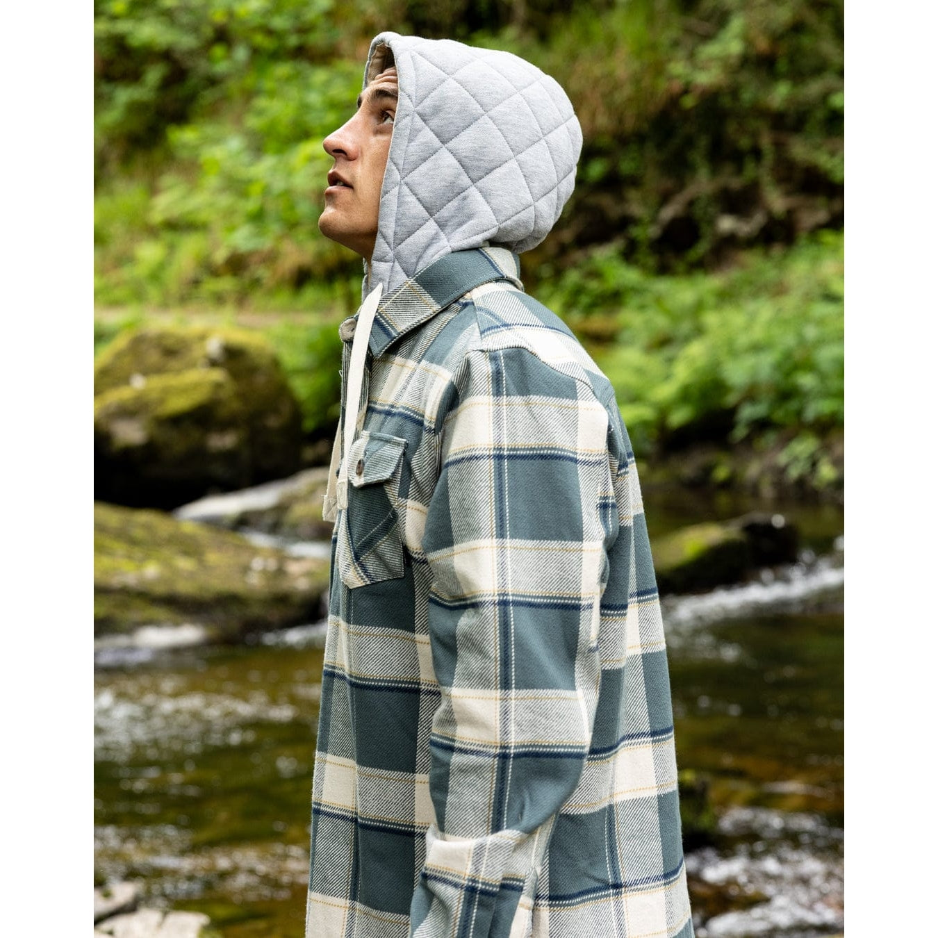 Side view of a man wearing a Saltrock Strait long sleeve check shirt in dark teal green with grey detachable hood outdoors