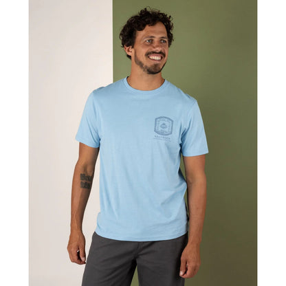 Man wearing a light blue t-shirt with a logo against a green wall