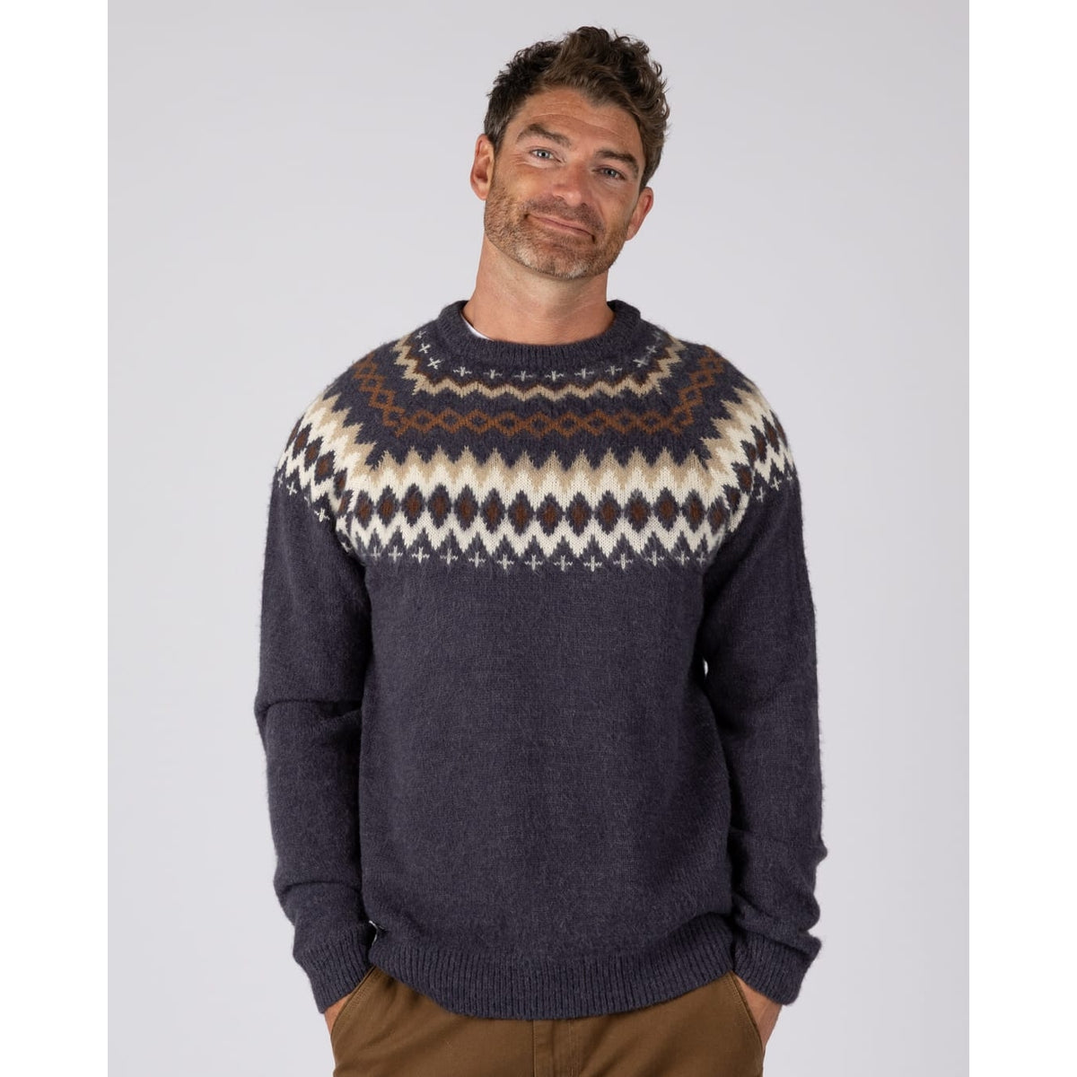 A man wearing a  Saltrock Fairisle Rock crew neck knitted jumper in blue with a geometric Fairisle style pattern across the chest and shoulders.