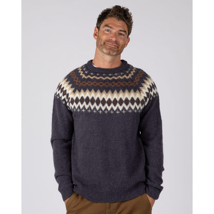 A man wearing a  Saltrock Fairisle Rock crew neck knitted jumper in blue with a geometric Fairisle style pattern across the chest and shoulders.