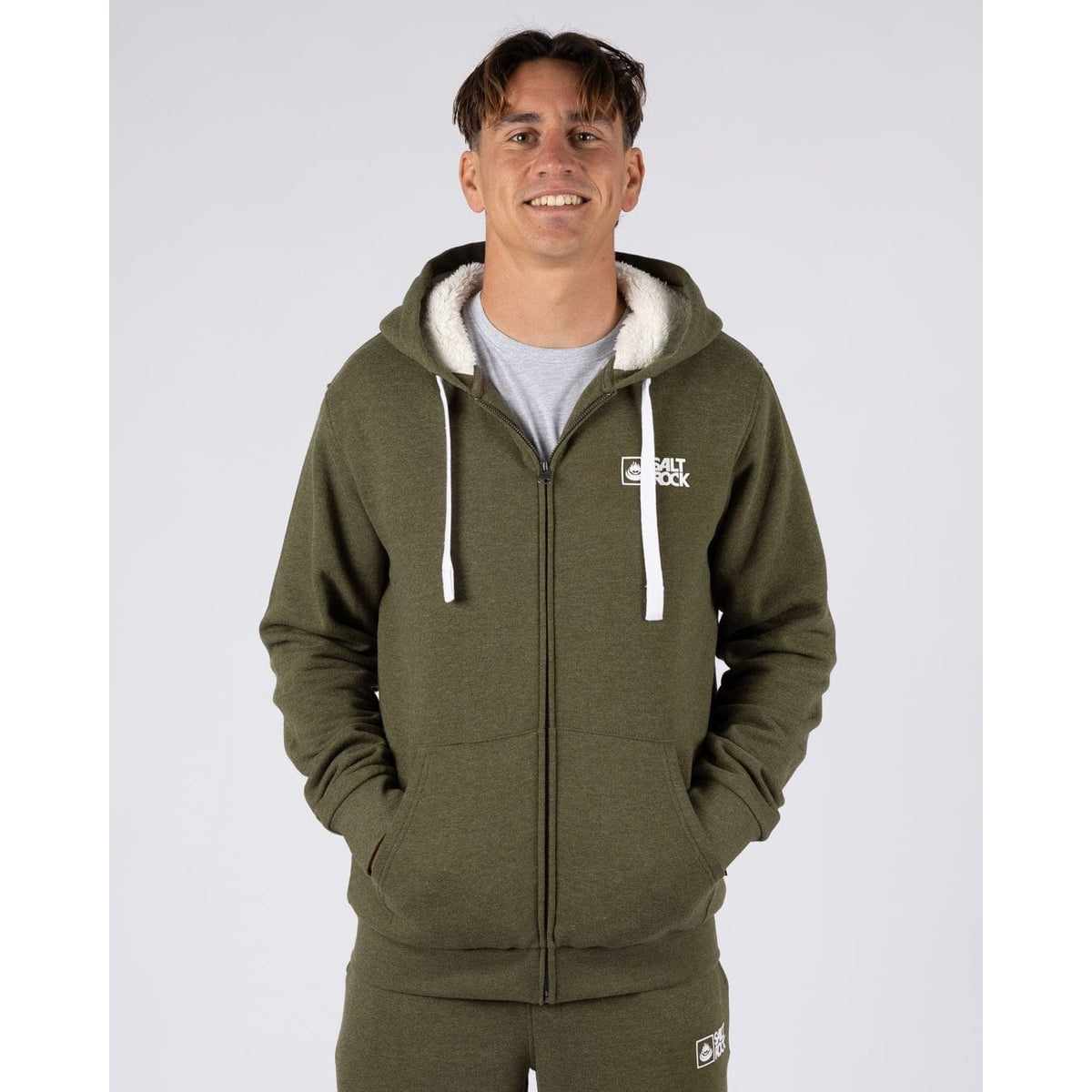 Front view of a man wearing a Saltrock Essential full zip faux fur lined hoodie in khaki green with his hands in the tummy pockets.