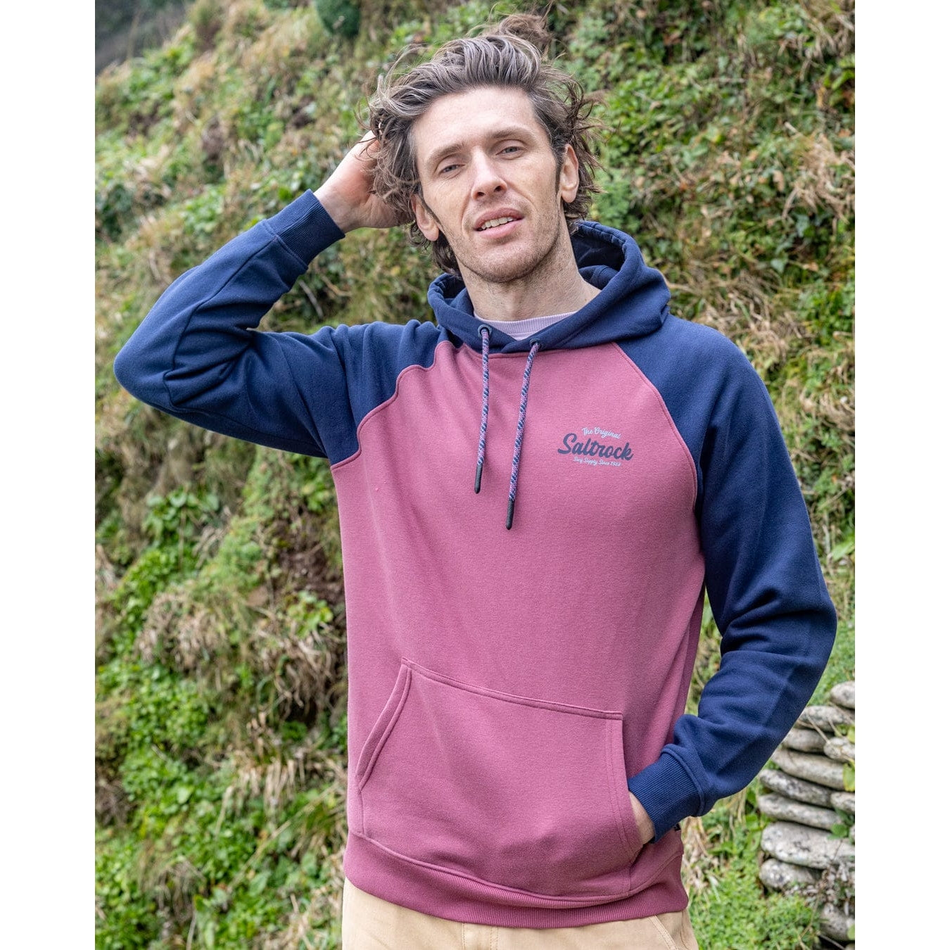 Front view of a man wearing a Saltrock Postcard Script raglan pop hoodie in dark pink with navy blue sleeves and hood stood outdoors