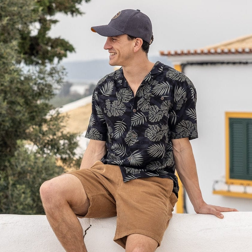 A man wearing a short sleeve Saltrock Warp Surf Resort Shirt in dark grey with a geometric and green tropical leaf pattern with tan shorts