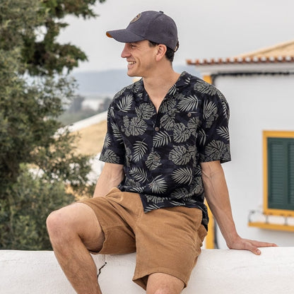 A man wearing a short sleeve Saltrock Warp Surf Resort Shirt in dark grey with a geometric and green tropical leaf pattern with tan shorts