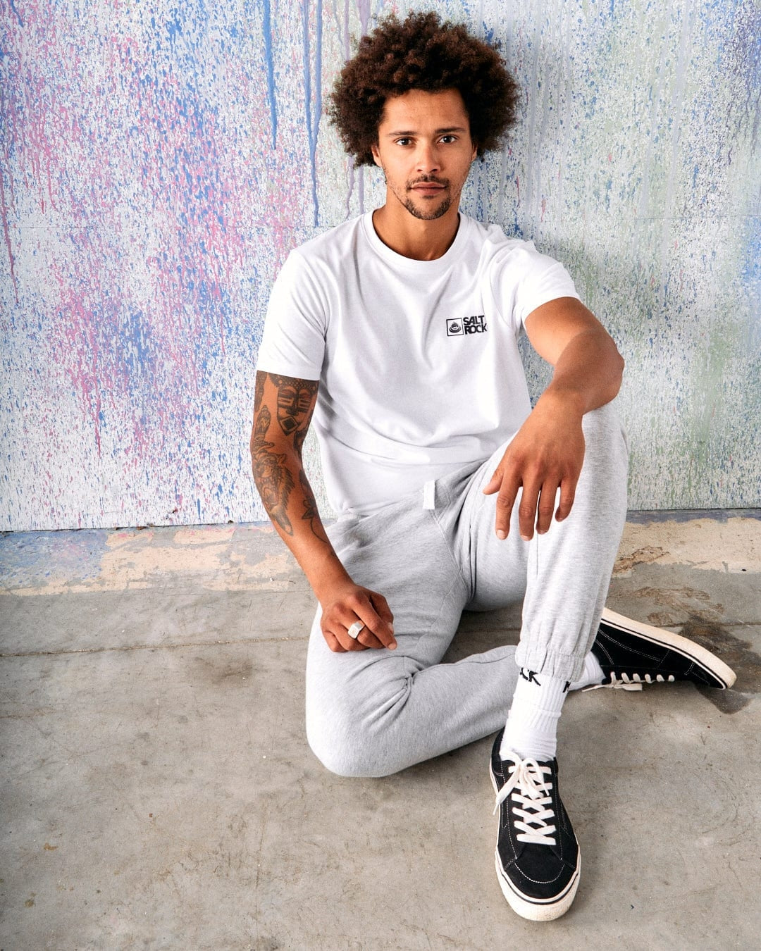 Man sat on the floor leaning against a wall wearing a pair of mens grey Saltrock jogger sweat trousers with a white t-shirt