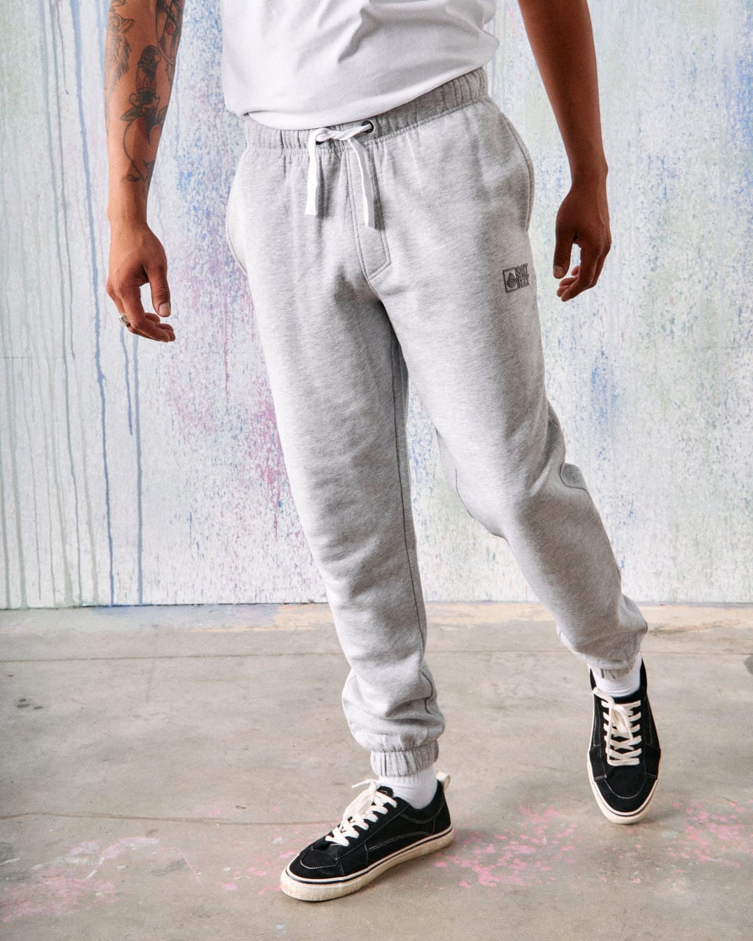 A man wearing a pair of Saltrock Original grey jogger sweat trousers and a white t-shirt standing in front of a colourful painted background.