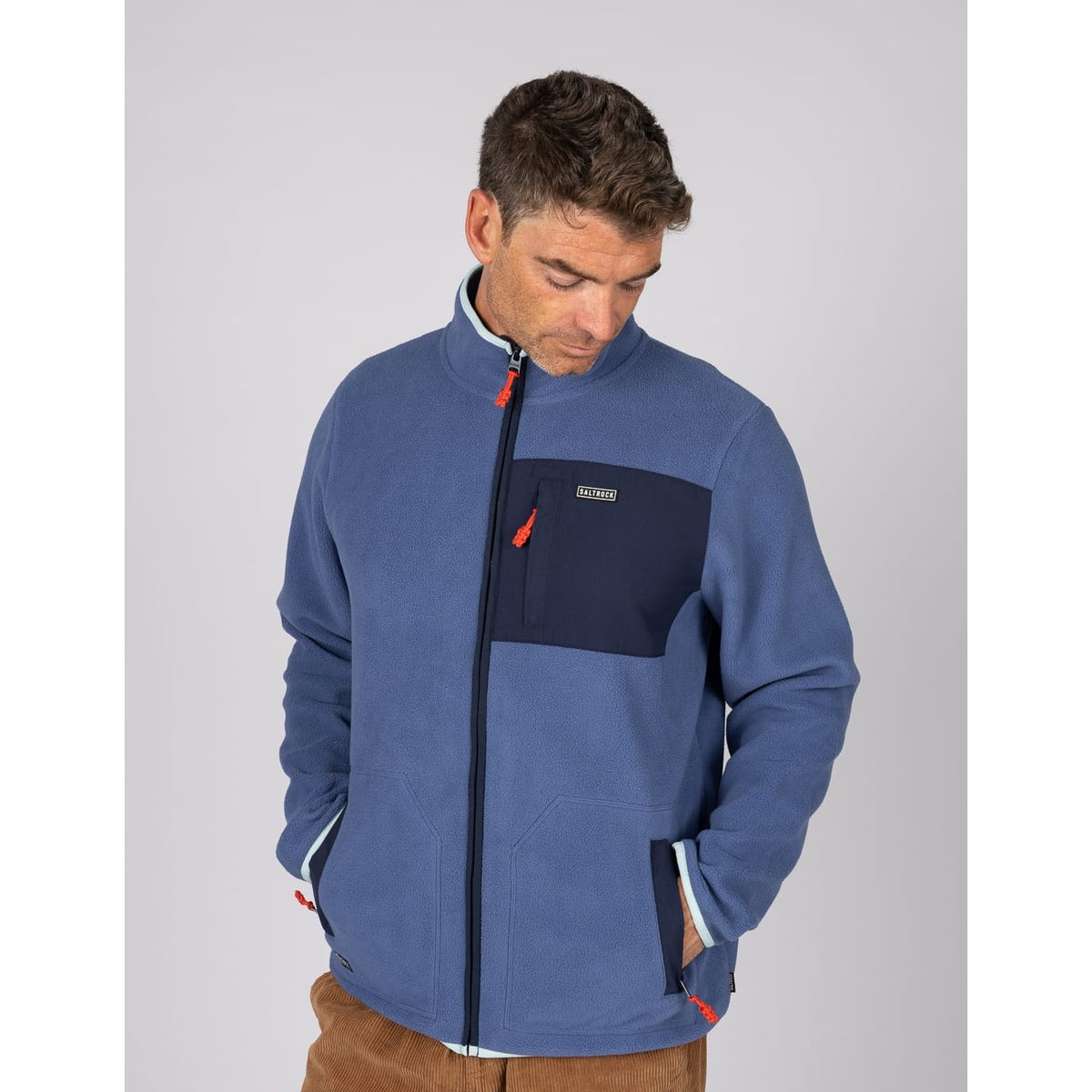 Front view of a man wearing a Saltrock full zip Leo fleece in blue, with contrasting navy, turquoise and red trim, tummy pockets and one zip pocket on the chest.