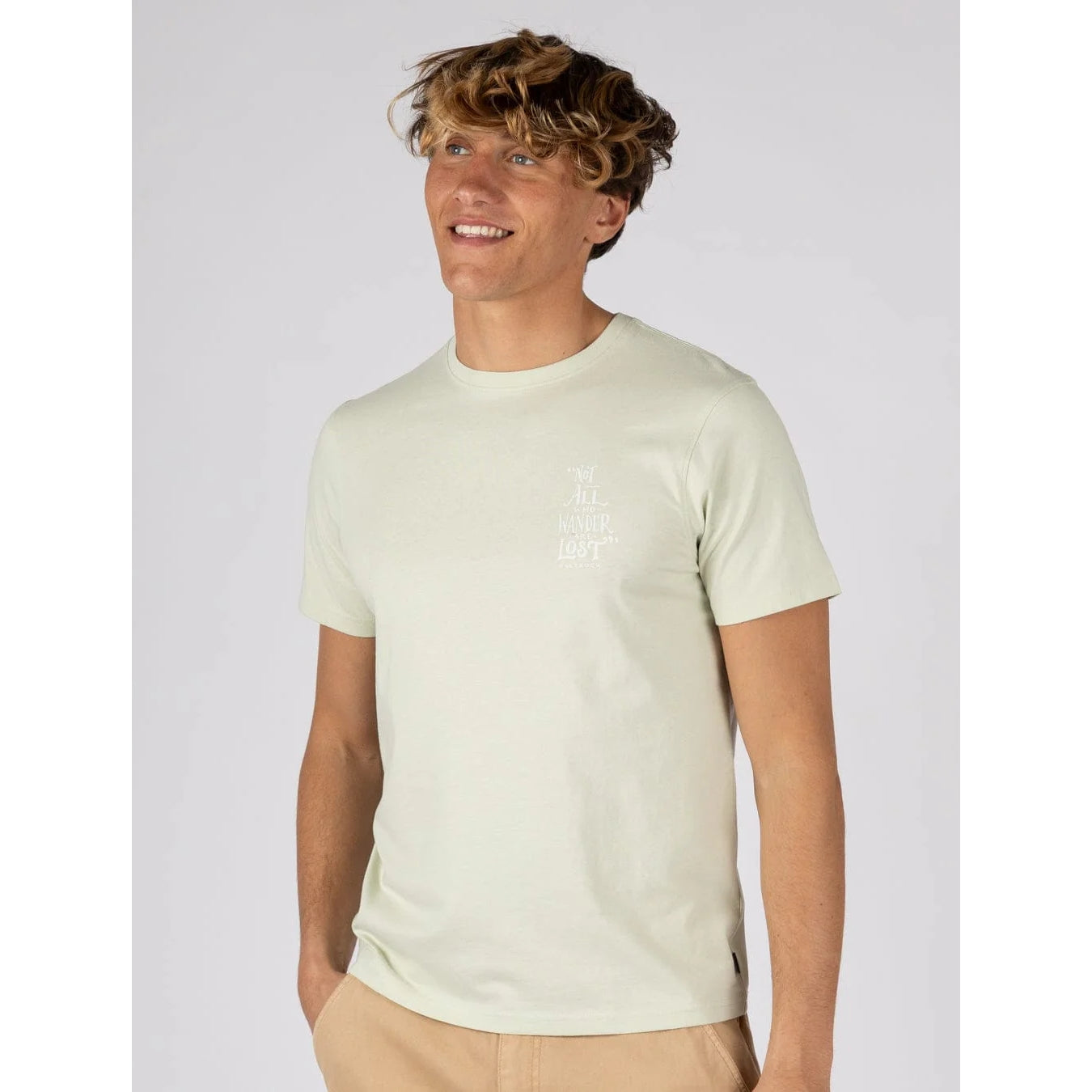 Front view of a man wearing a Saltrock short sleeve Lost Ships t-shirt in light green featuring a small print of the phrase "Not all who Wander are Lost"