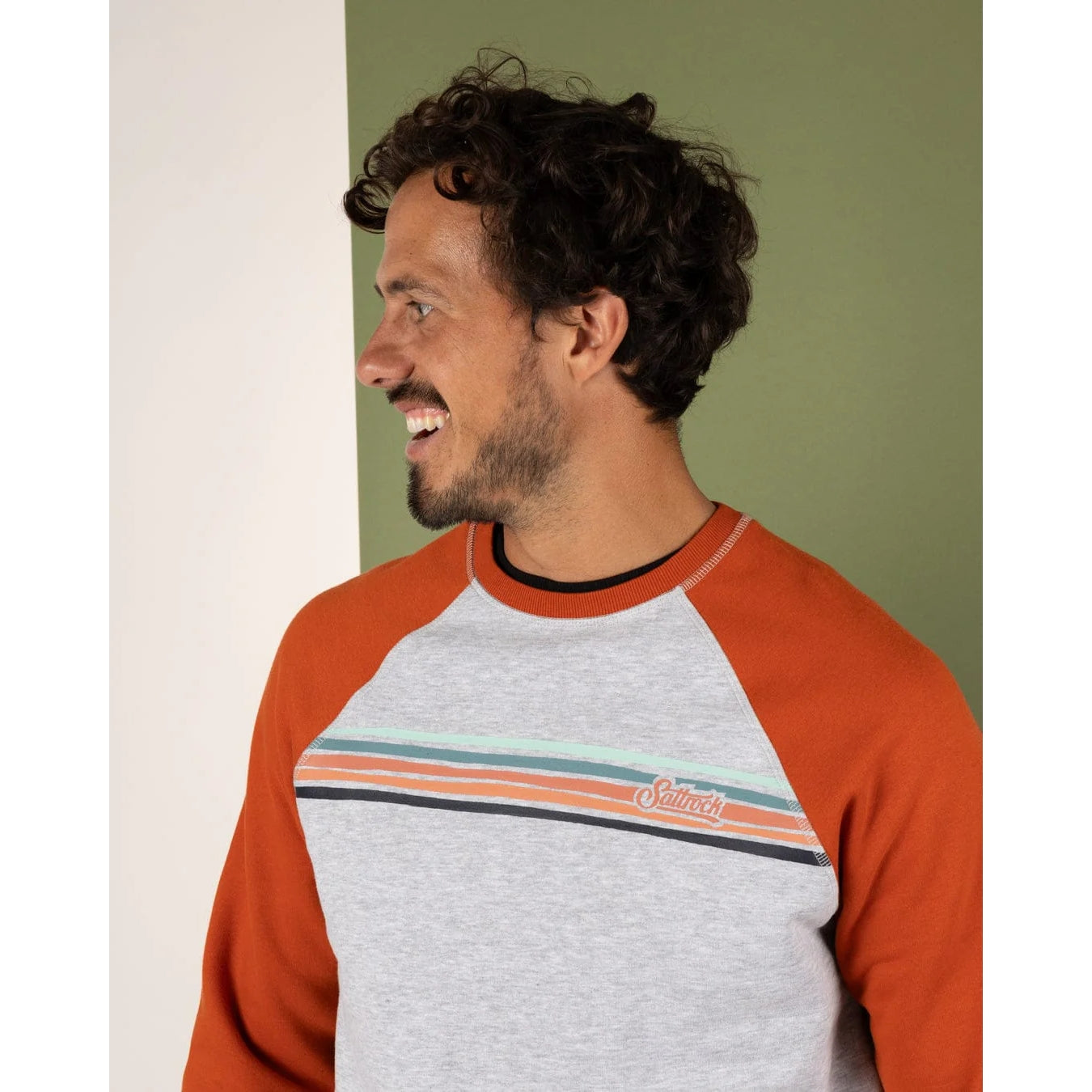 Man wearing a grey and orange long-sleeve shirt with a brand logo on a green wall.