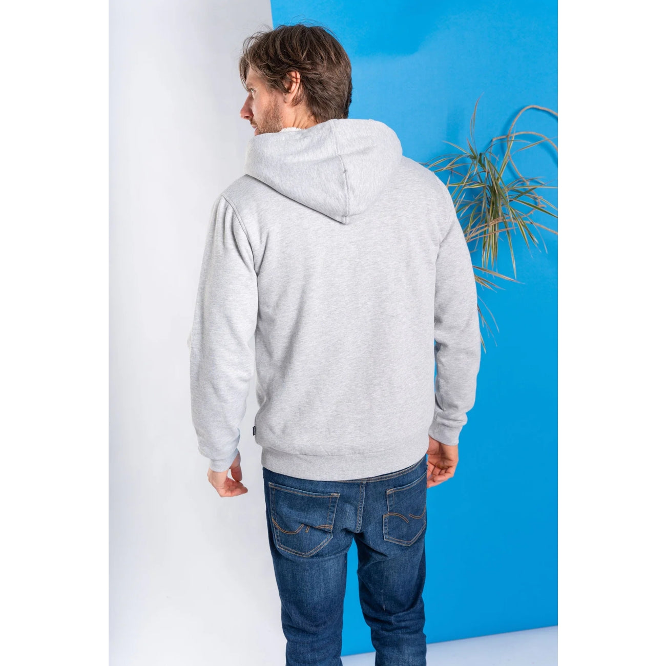 Man wearing a grey Saltrock faux fur lined Original zip hoodie and blue jeans against a white and blue wall.