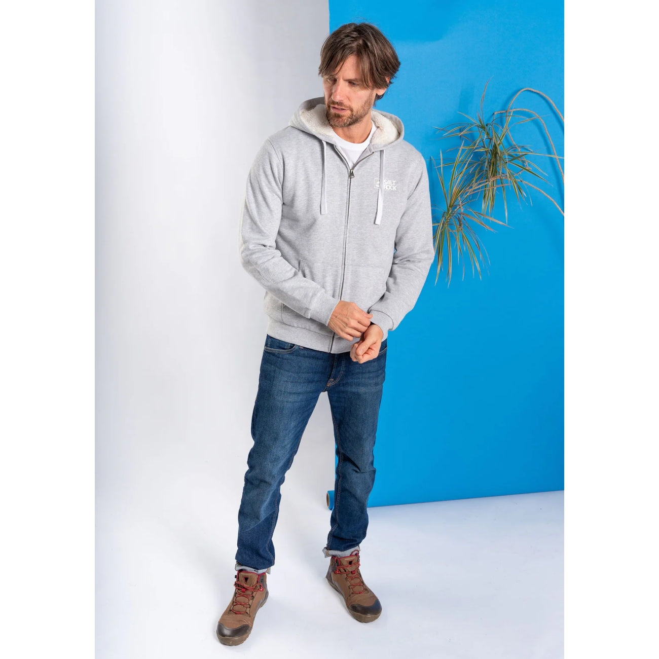 Man wearing a grey Saltrock Original faux fur lined hoodie and blue jeans standing against a white and blue background.
