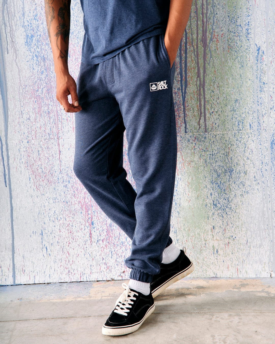 Front view of a man wearing dark blue Saltrock Original jogger sweat trousers and sneakers, standing in front of a colourful background.