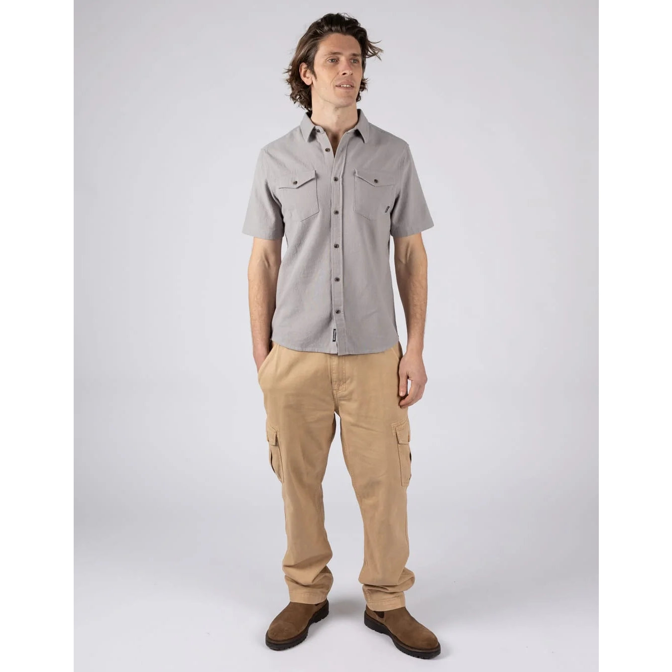 A man wearing a Saltrock short sleeve Polperro button shirt in grey with a waffle texture and two button pockets on the chest with tan trousers and brown boots.