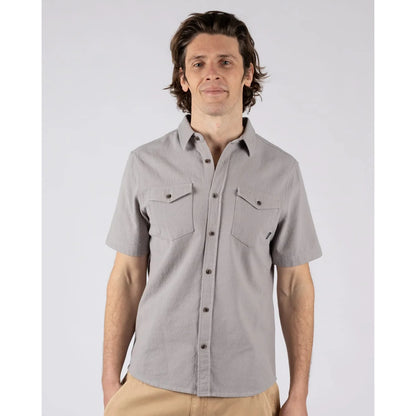 A man wearing a Saltrock short sleeve Polperro button shirt in grey with a waffle texture and two button pockets on the chest with tan trousers.