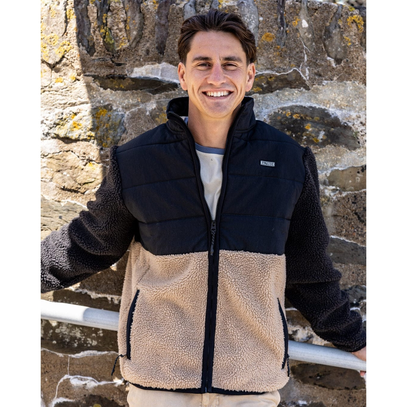 Man wearing a two-tone Saltrock Rafe fleece jacket in black and beige standing against a stone wall.