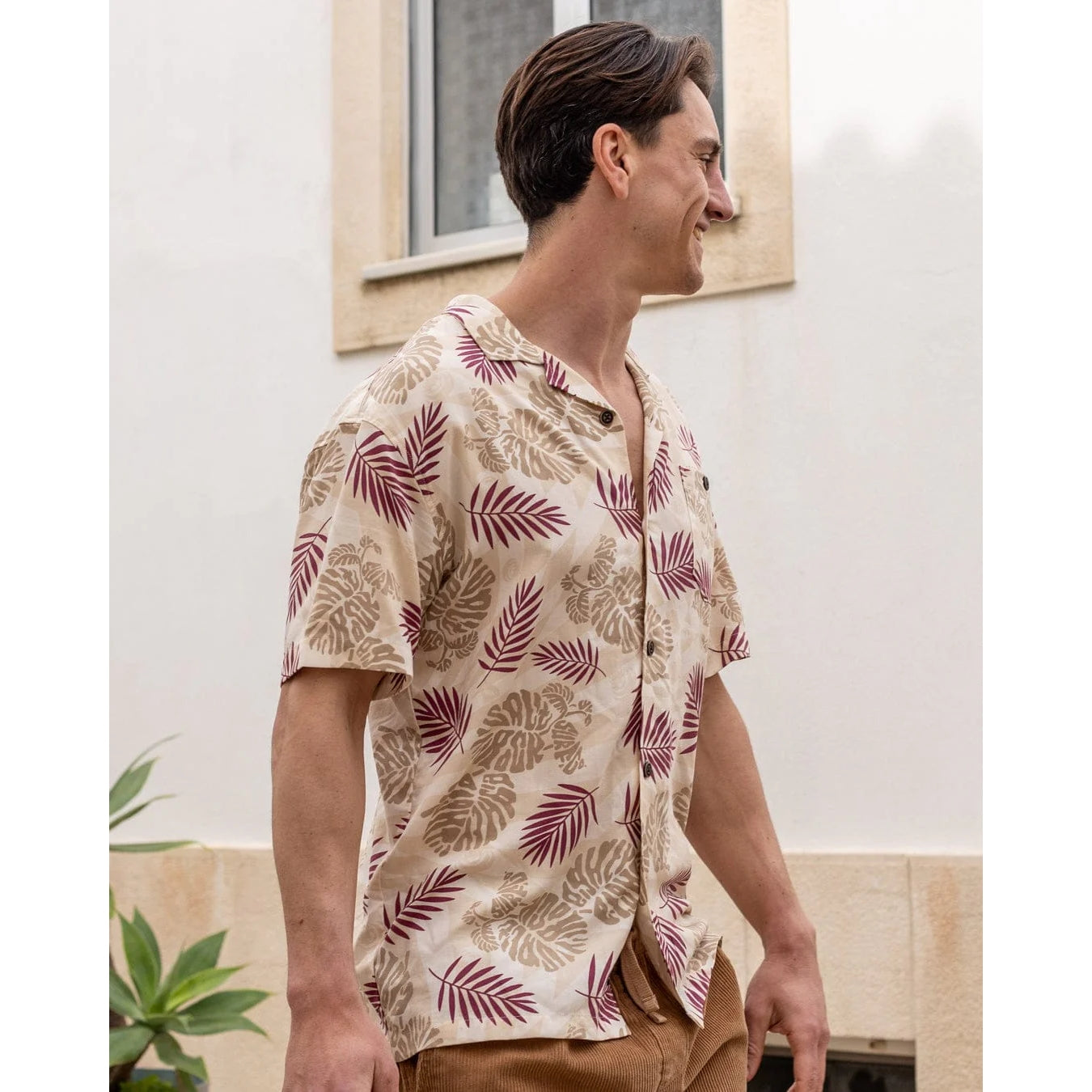 Side view of a man wearing a Saltrock short sleeve Warp Surf Resort shirt in cream with a dark red and tan tropical leaves print with tan shorts outdoors