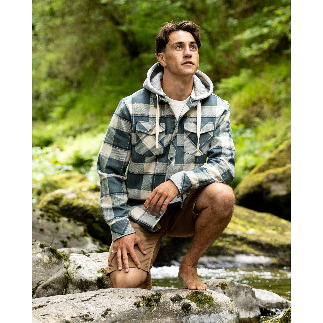 Man wearing a Saltrock Strait long sleeve check shirt in dark teal green with chest pockets, detachable hood and tan shorts in an outdoor setting