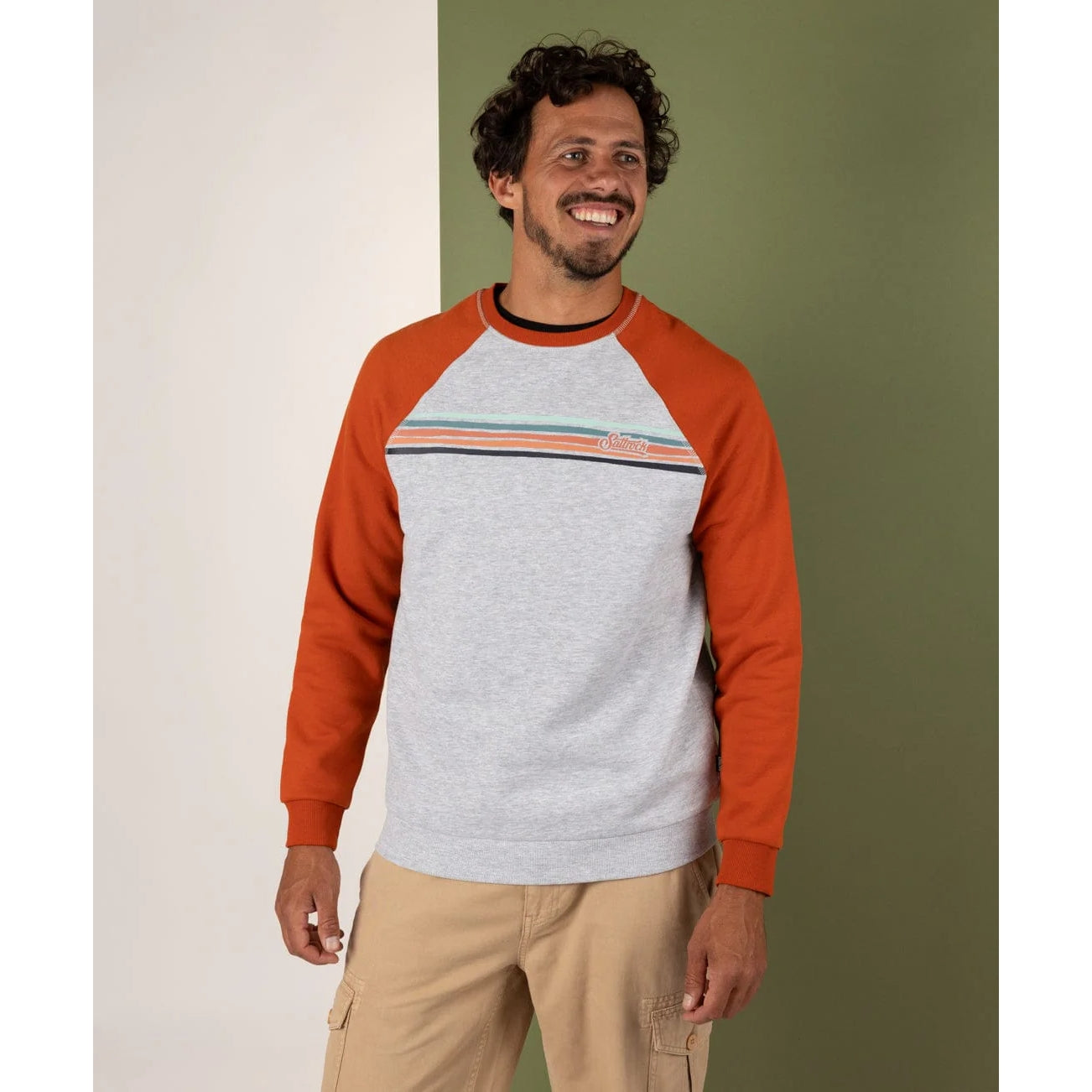 Man wearing a grey and orange sweatshirt with beige pants against a green wall.