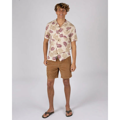 A man wearing a Saltrock short sleeve Warp Surf Resort shirt in cream with a dark red and tan tropical leaves print with tan shorts and flip flops