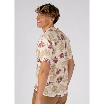 Side view of a man wearing a Saltrock short sleeve Warp Surf Resort shirt in cream with a dark red and tan tropical leaves print with tan shorts