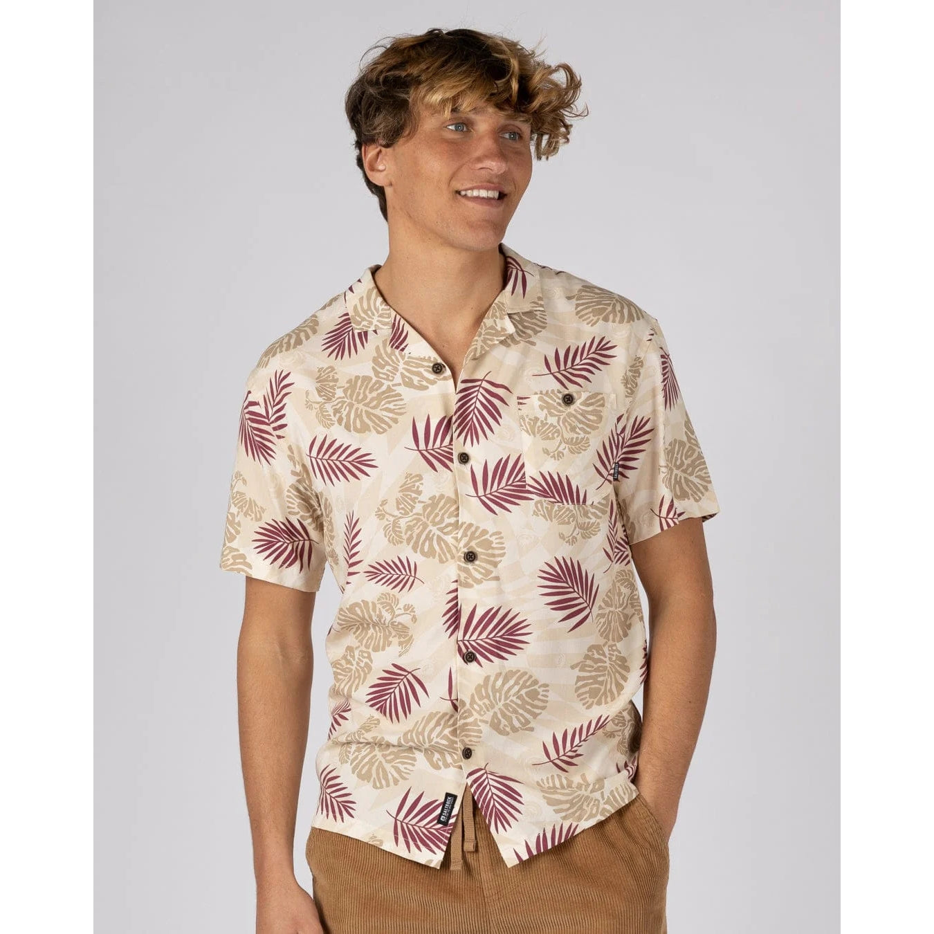 A man wearing a Saltrock short sleeve Warp Surf Resort shirt in cream with a dark red and tan tropical leaves print with tan shorts