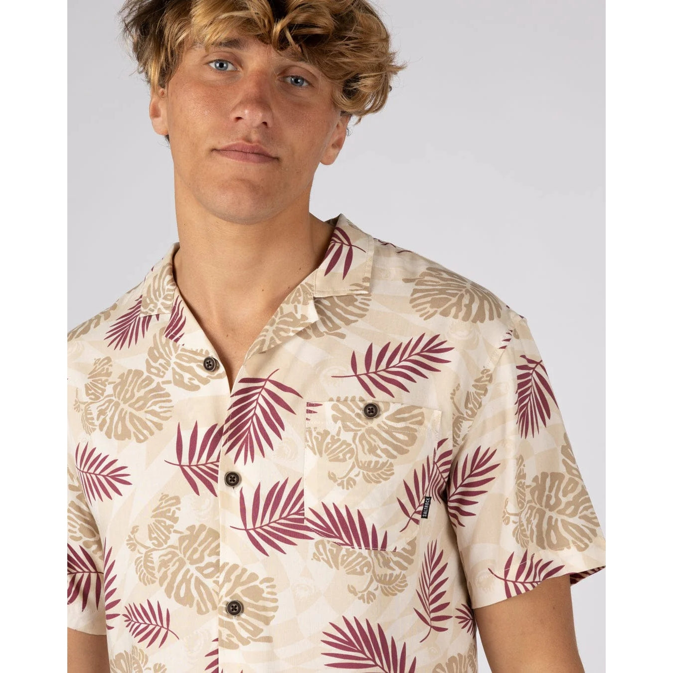 Close-up view of a man wearing a Saltrock short sleeve Warp Surf Resort shirt in cream with a dark red and tan tropical leaves with a chest pocket