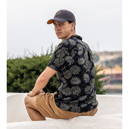 Back view of a man sat outside wearing a short sleeve Saltrock Warp Surf Resort Shirt in dark grey with a geometric and green tropical leaf print with tan shorts