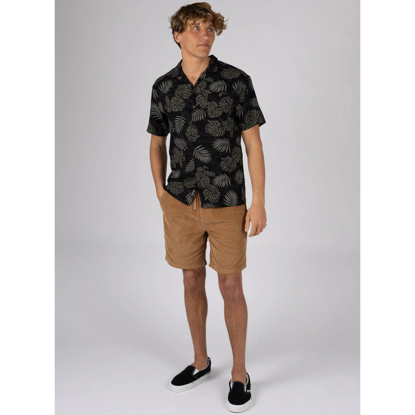 Man wearing a short sleeve Saltrock Warp Surf Resort Shirt in dark grey with a geometric and green tropical leaf print, with tan shorts and navy slip on shoes