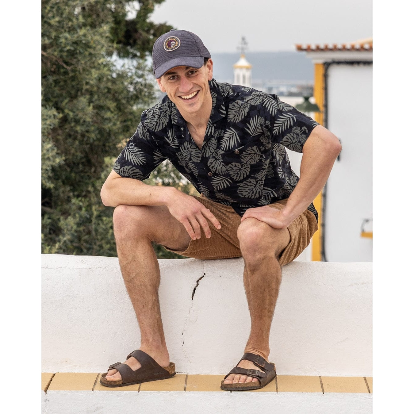 Man wearing a short sleeve Saltrock Warp Surf Resort Shirt in dark grey with a geometric and green tropical leaf print, with shorts and brown sandals outdoors