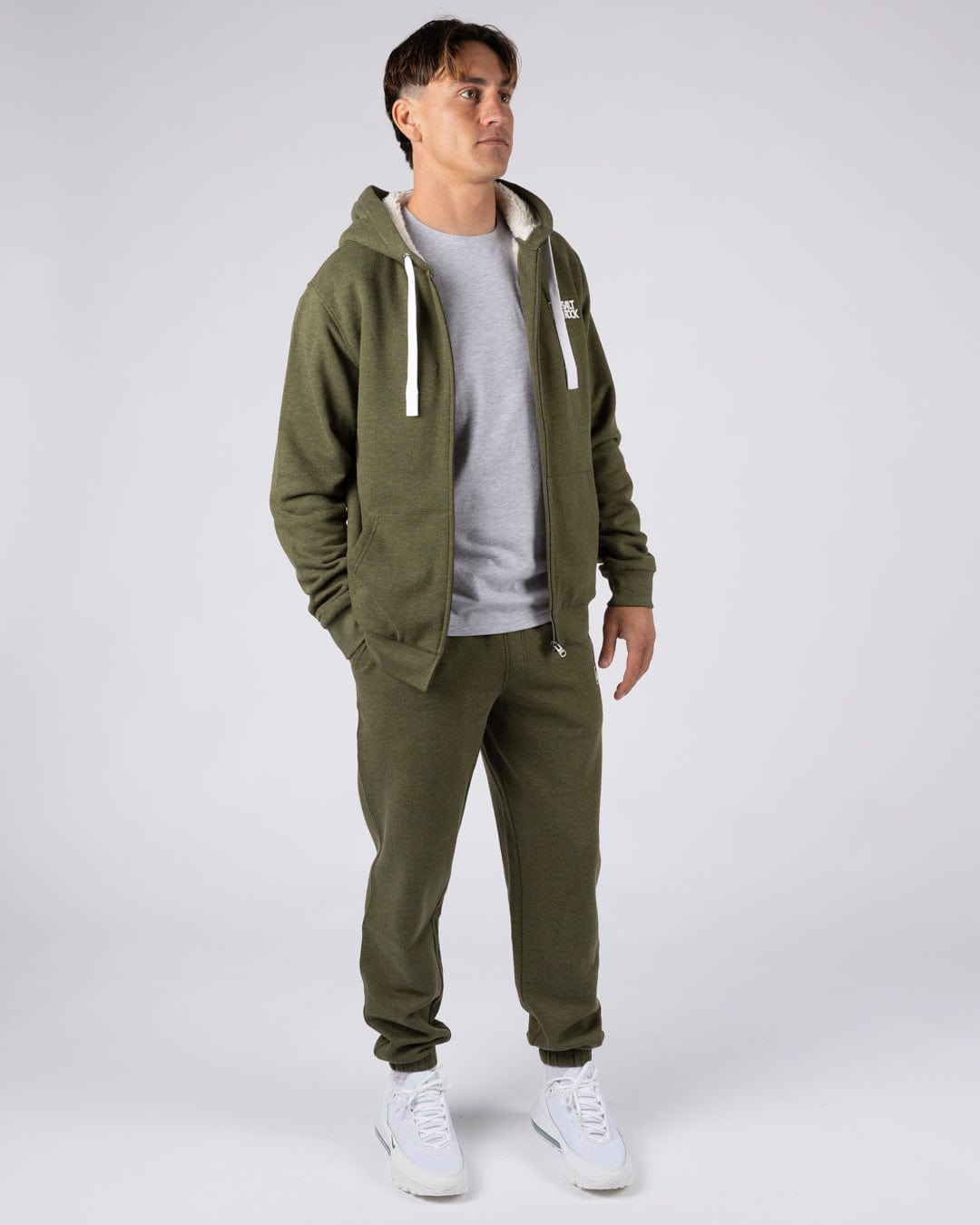 A man wearing a Saltrock Essential full zip faux fur lined hoodie in khaki green with khaki green trousers and white trainers.