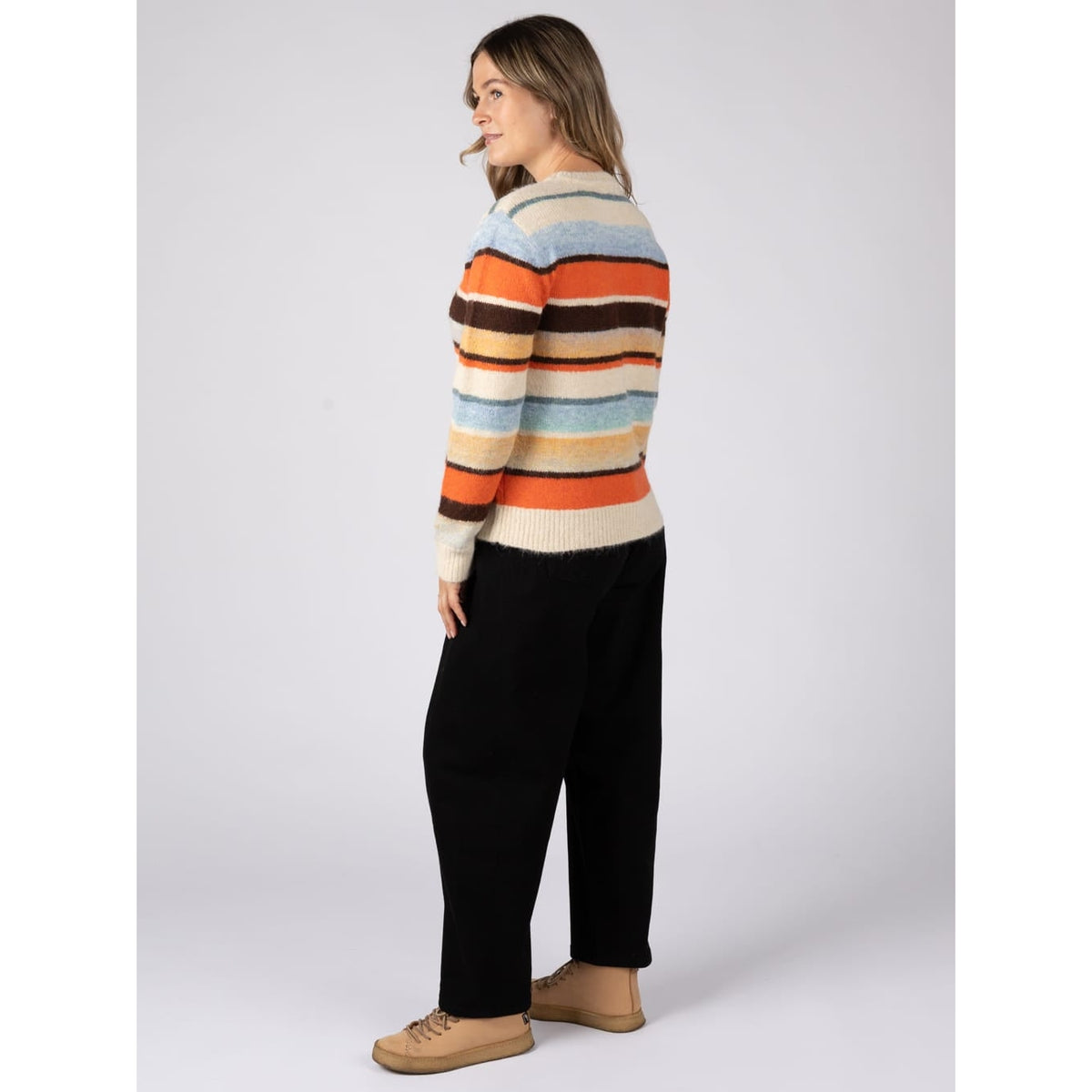 Back view of a woman wearing a Saltrock Lyris crew neck knitted jumper in an orange, cream and multicolour striped pattern with black trousers.
