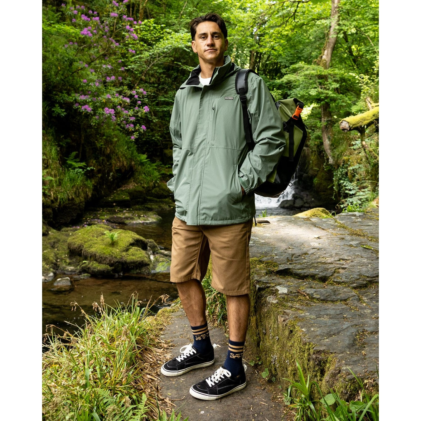 Front view of a man wearing a Saltrock Whistler II waterproof hooded jacket in light green with tan shorts and trainers outdoors