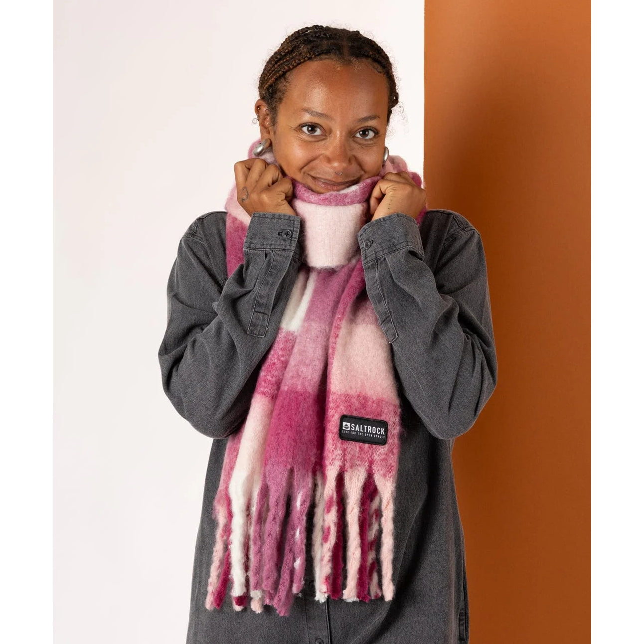 Woman wearing a Saltrock Harvest pink check scarf with tassels and a brand logo against a white and orange background