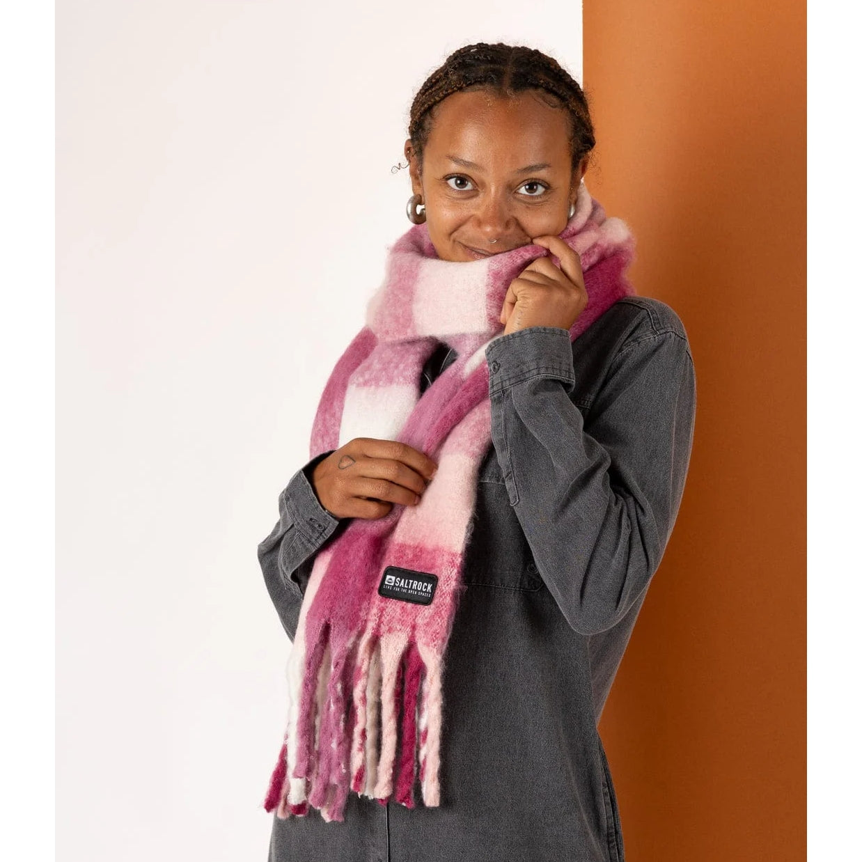 Woman wearing a pink check Saltrock Harvest scarf with a brand logo against a white and orange background