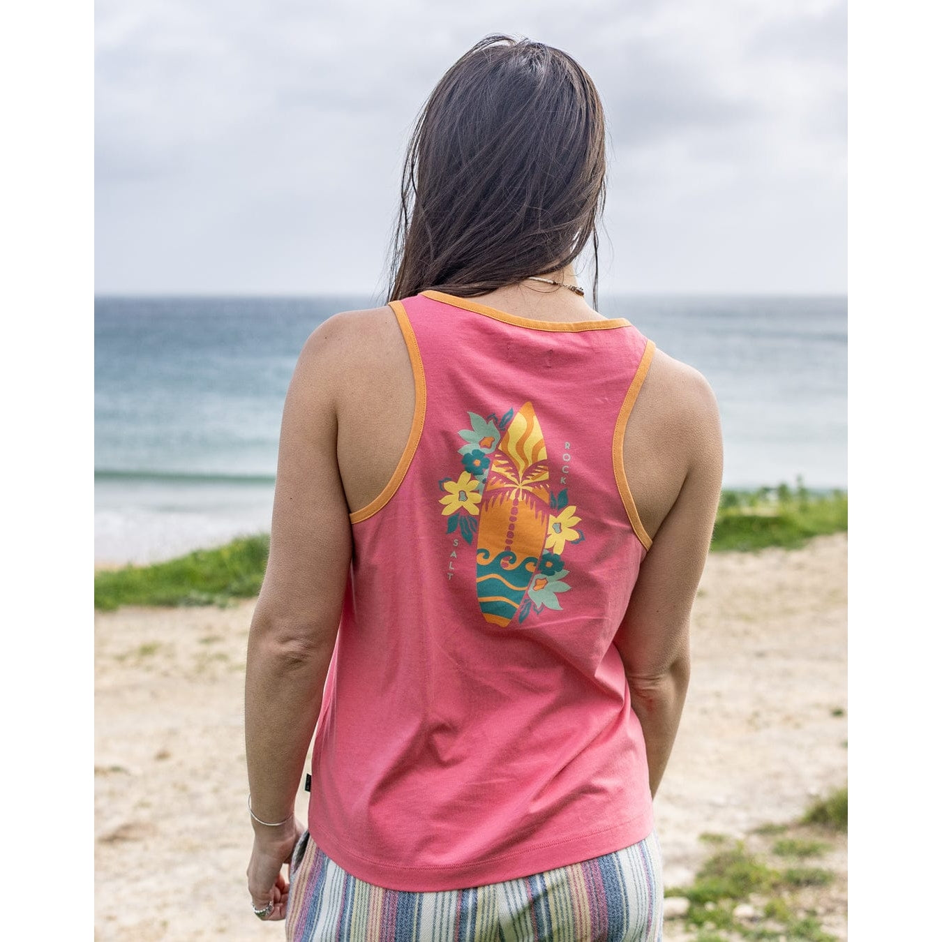 Back view of a woman wearing a Saltrock pink Sun Surfer vest top with a colourful floral surfboard print on a beach