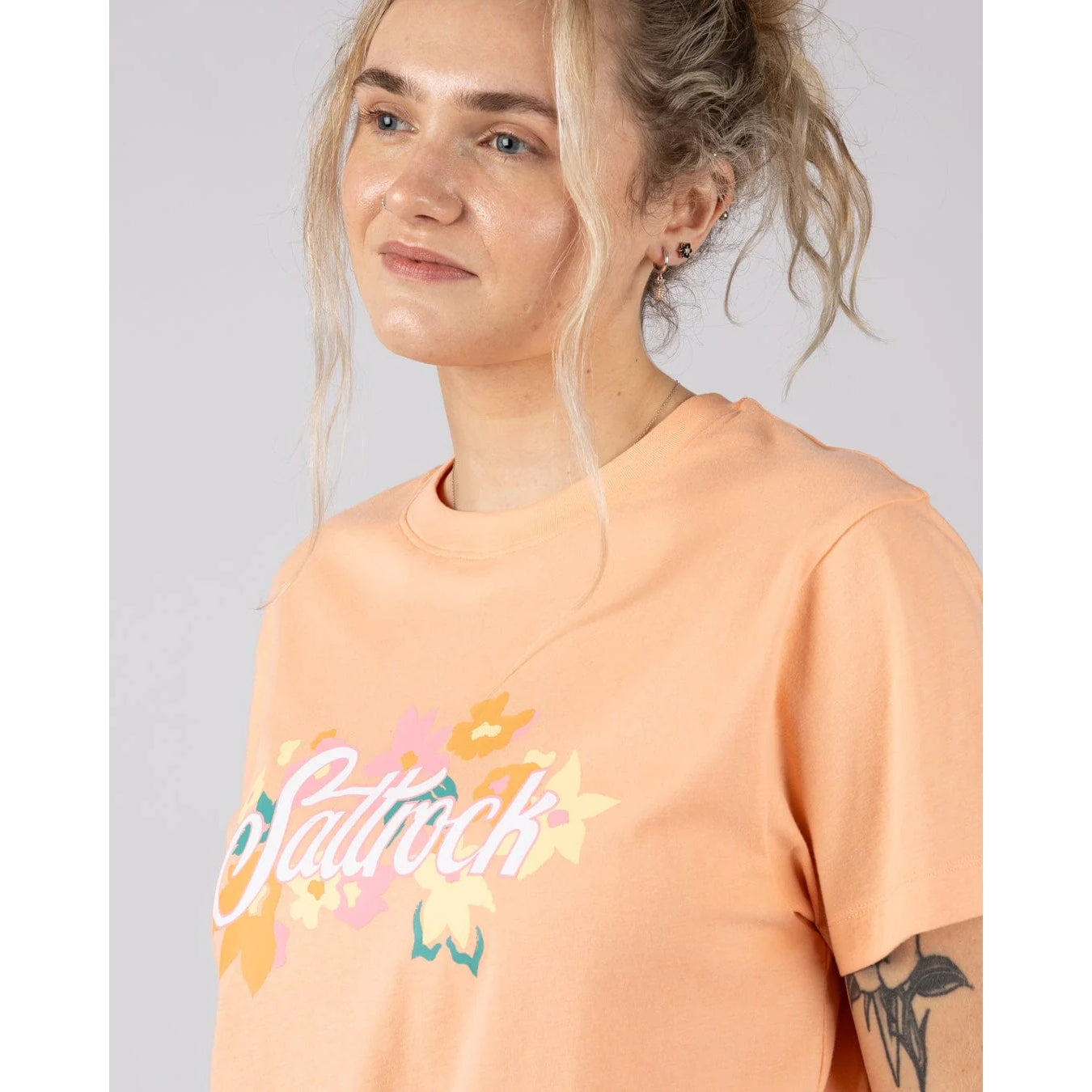 Close-up view of a woman wearing a peach-coloured Saltrock short sleeve Trader Daze t-shirt with colourful floral logo on the chest