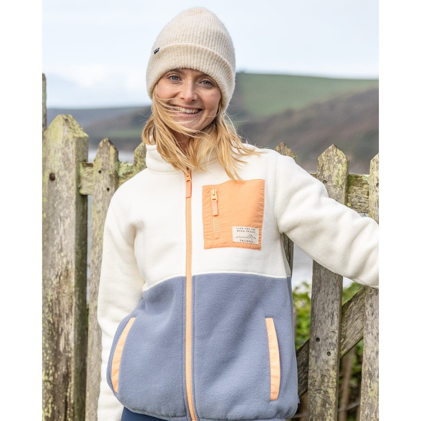 Front view of a woman wearing a Saltrock Denya full zip colour block fleece in cream, blue and coral, against a wooden fence and scenic background