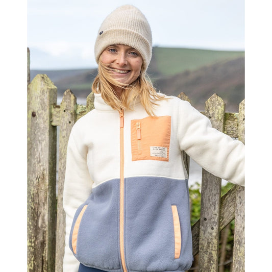 Front view of a woman wearing a Saltrock Denya full zip colour block fleece in cream, blue and coral, against a wooden fence and scenic background