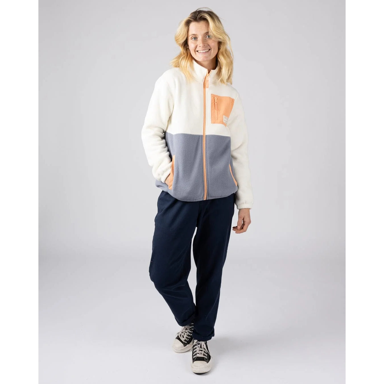 Woman wearing a Saltrock full zip Denya fleece with a colour-block pattern in cream, blue and coral with navy pants on a grey background