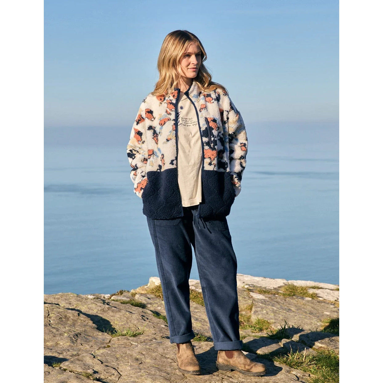 Woman wearing a patterned Saltrock Folie fleece and dark trousers standing on a rocky outcrop with ocean view