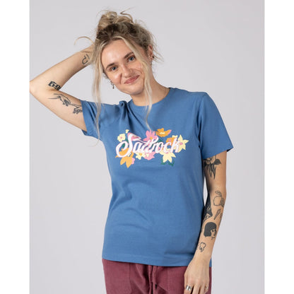 Front view of a woman wearing a bright blue Saltrock Trader Daze t-shirt with floral logo print on the chest with burgundy trousers on a plain background