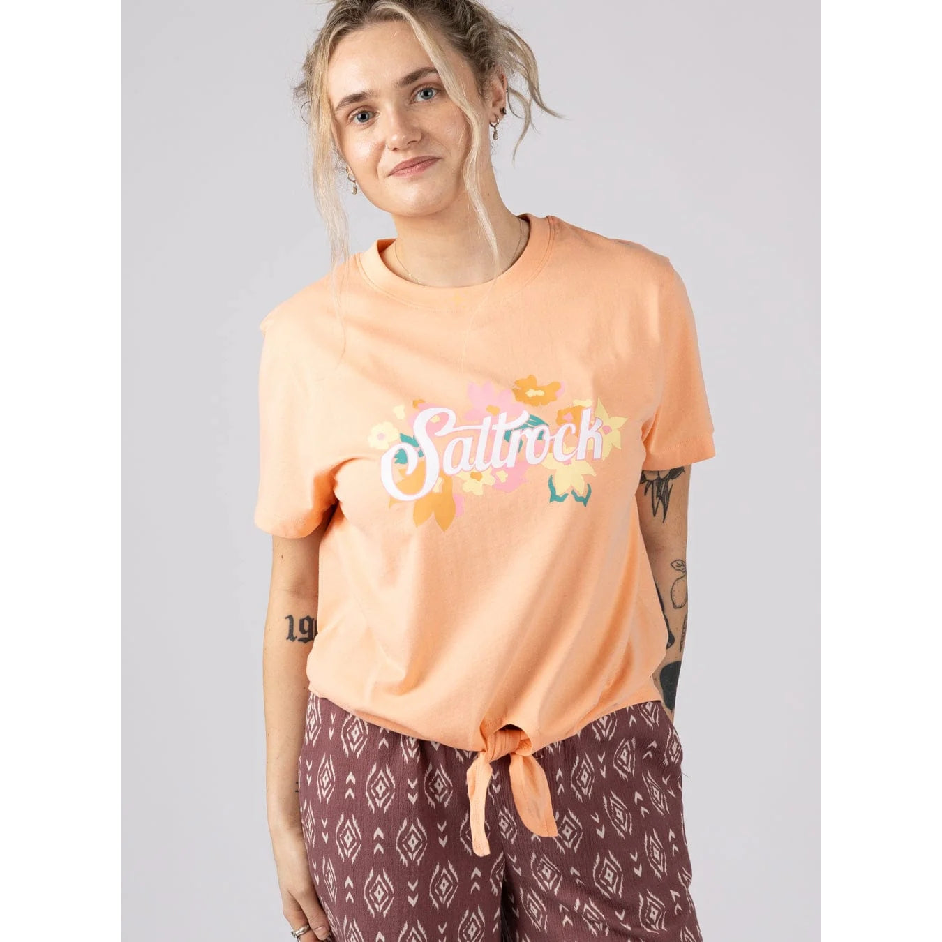 Front view of a woman wearing a Saltrock short sleeve Trader Daze t-shirt in peach, with logo print on the chest and a knot tie hem.