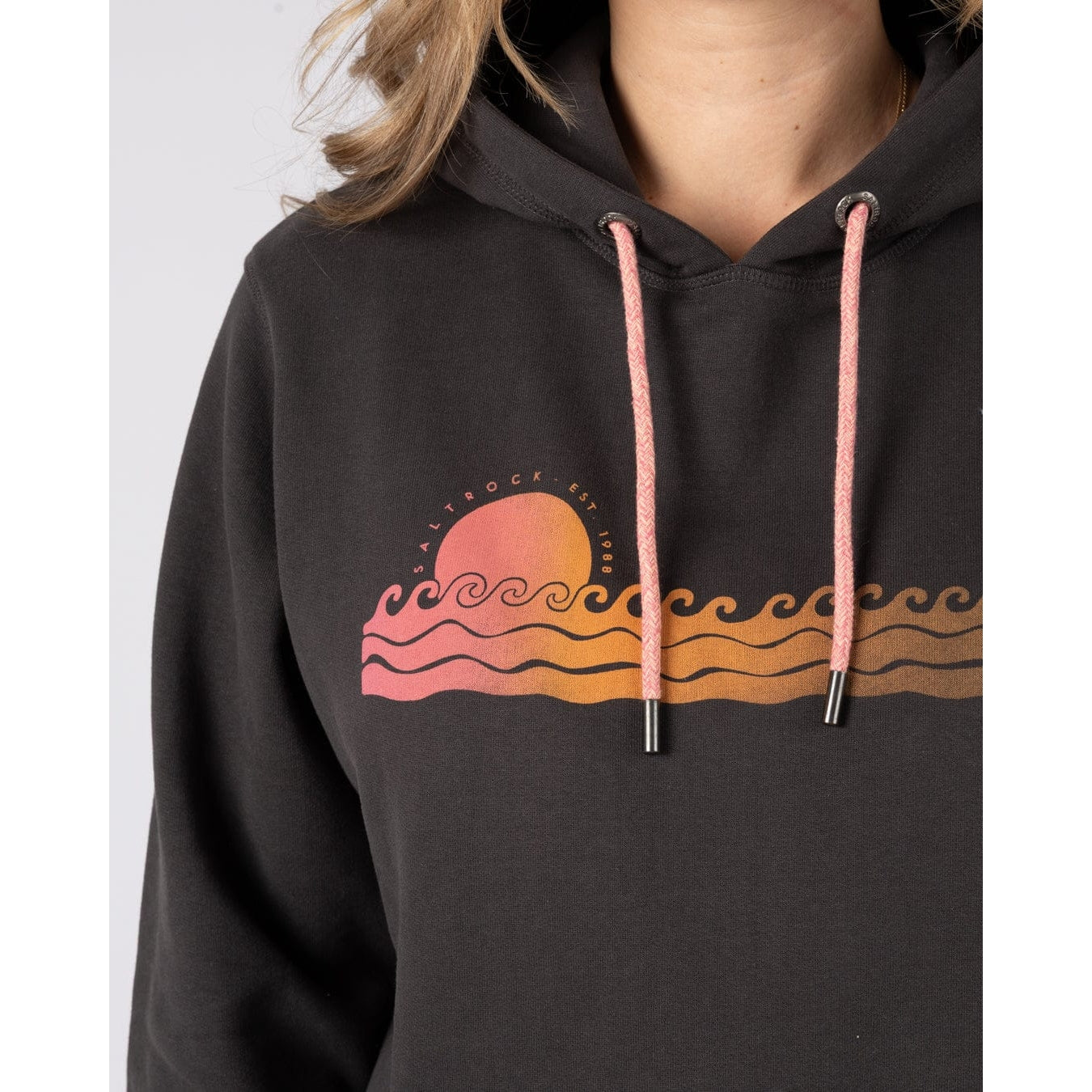Close-up view of a woman wearing a Saltrock Golden Hour pop hoodie with a wave and sun chest print on a white background