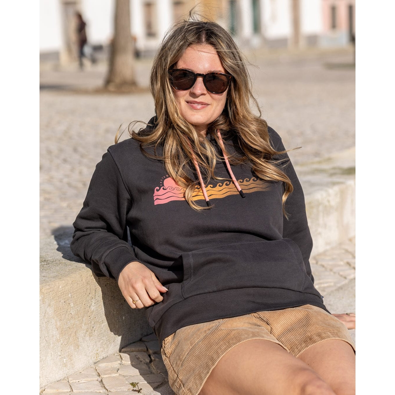 Woman wearing a Saltrock dark grey Golden Hour print pop hoodie with beige shorts, sitting outdoors.