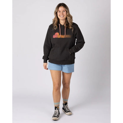 Woman wearing a dark grey Saltrock Golden Hour pop hoodie with a sun and sea chest print, blue shorts, and black sneakers on a white background