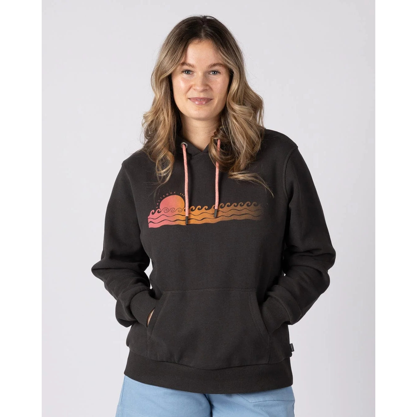 Front view of a woman wearing a dark grey Saltrock Golden Hour pop hoodie with a sun and sea chest print on a white background