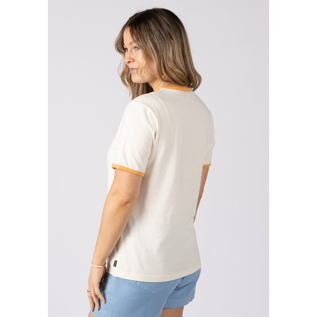 Side view of a woman wearing a Saltrock short sleeve Golden Hour t-shirt with orange trim and blue shorts on a plain background