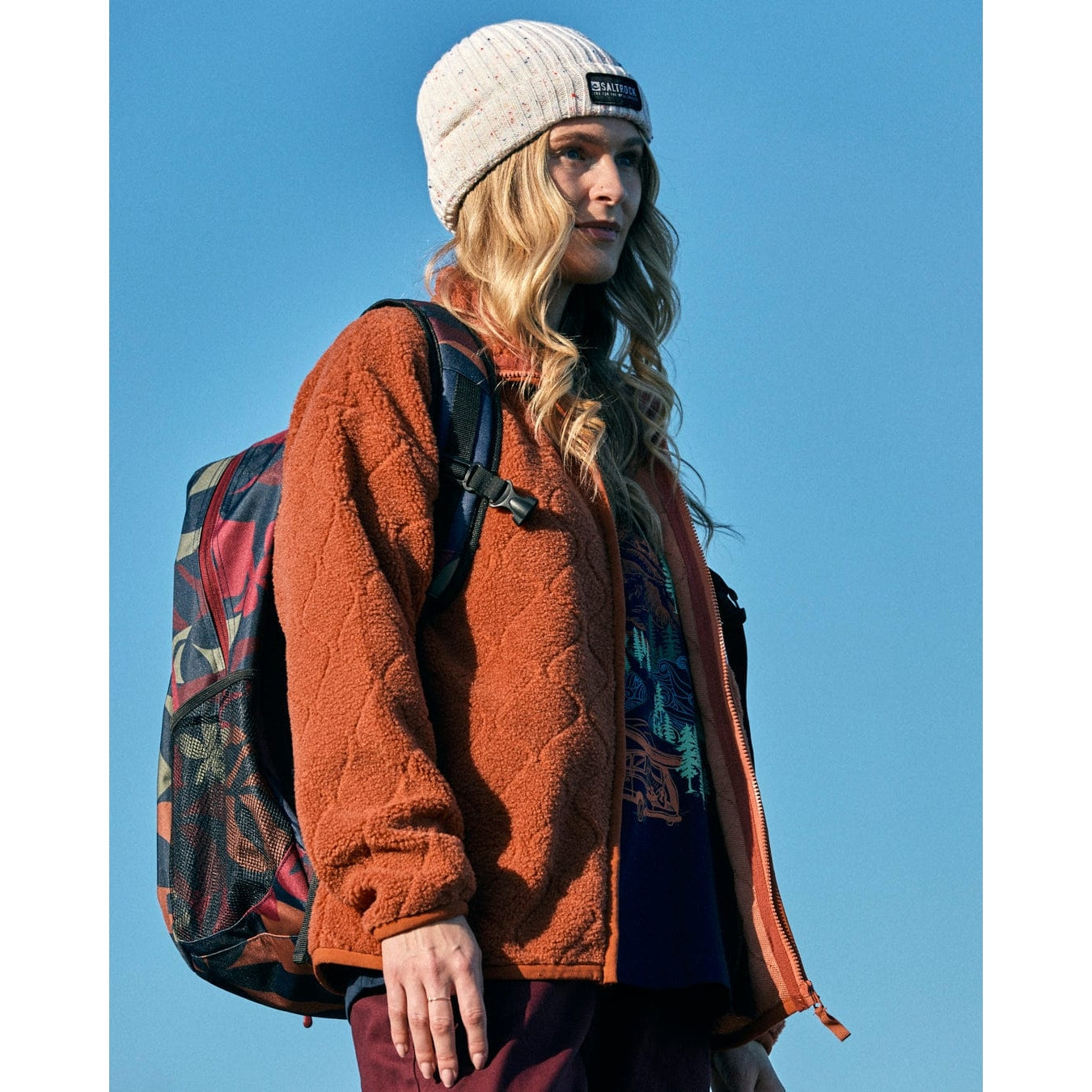 Woman wearing a Saltrock orange fleece jacket and white beanie with a backpack against a blue sky.