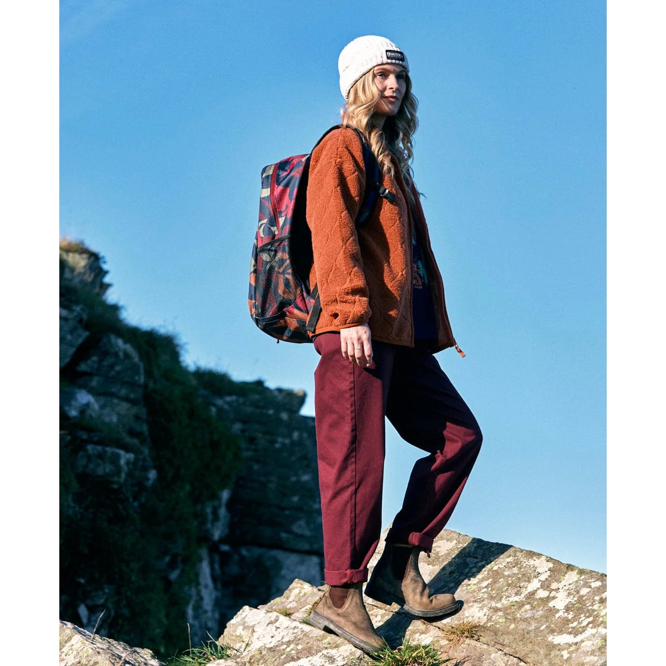 Woman wearing a Saltrock orange fleece ,with a backpack standing on a rocky outcrop against a blue sky